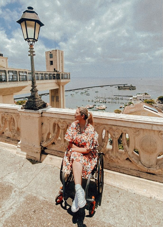 mulher na cadeira de rodas, de pernas cruzadas. Ao fundo o mar e o elevador lacerda em Salvador, na Bahia, Brasil