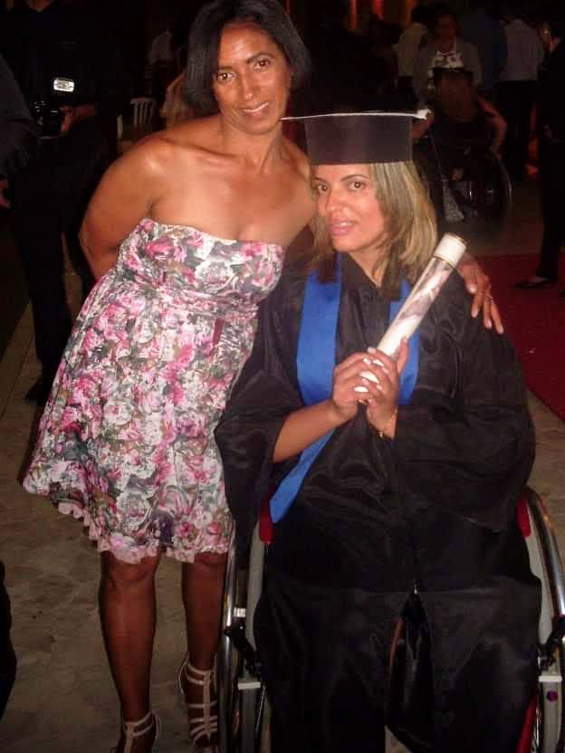 pessoas com deficiencia - mulher em cadeira de rodas, que usa uma beca e segura um diploma de formatura do ensino superior. Ao seu lado está uma mulher que a abraça
