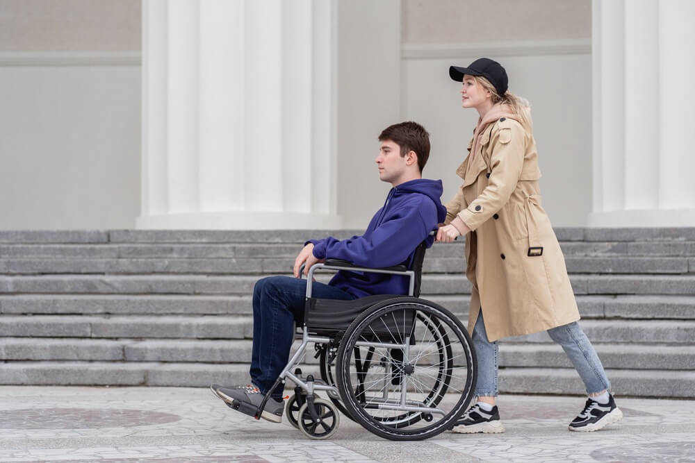 pessoas com deficiencia: homem em cadeira de rodas manual sendo conduzido por uma mulher.