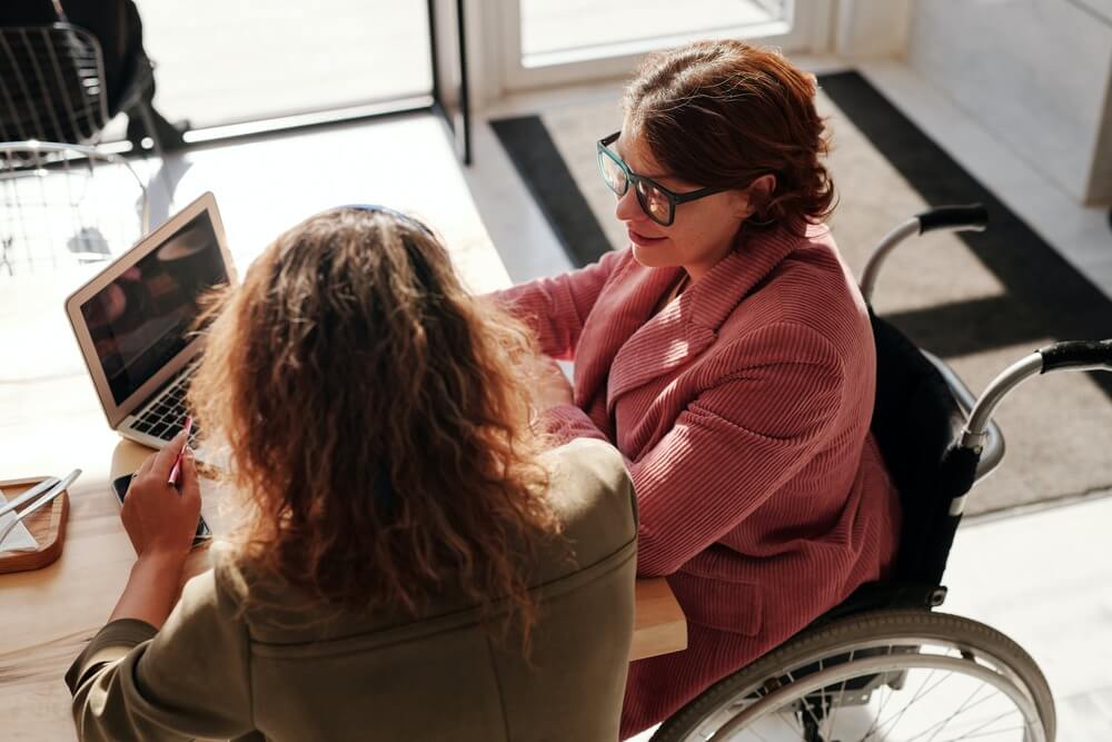 duas mulheres interagindo em um escritório, uma delas utiliza cadeira de rodas. Pessoas com deficiencia