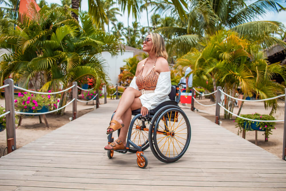 mulher cadeirante em esteira de madeira (deck) na praia. Ao fundo e ao seu redor grande vegetação e areia. Pessoas com deficiencia